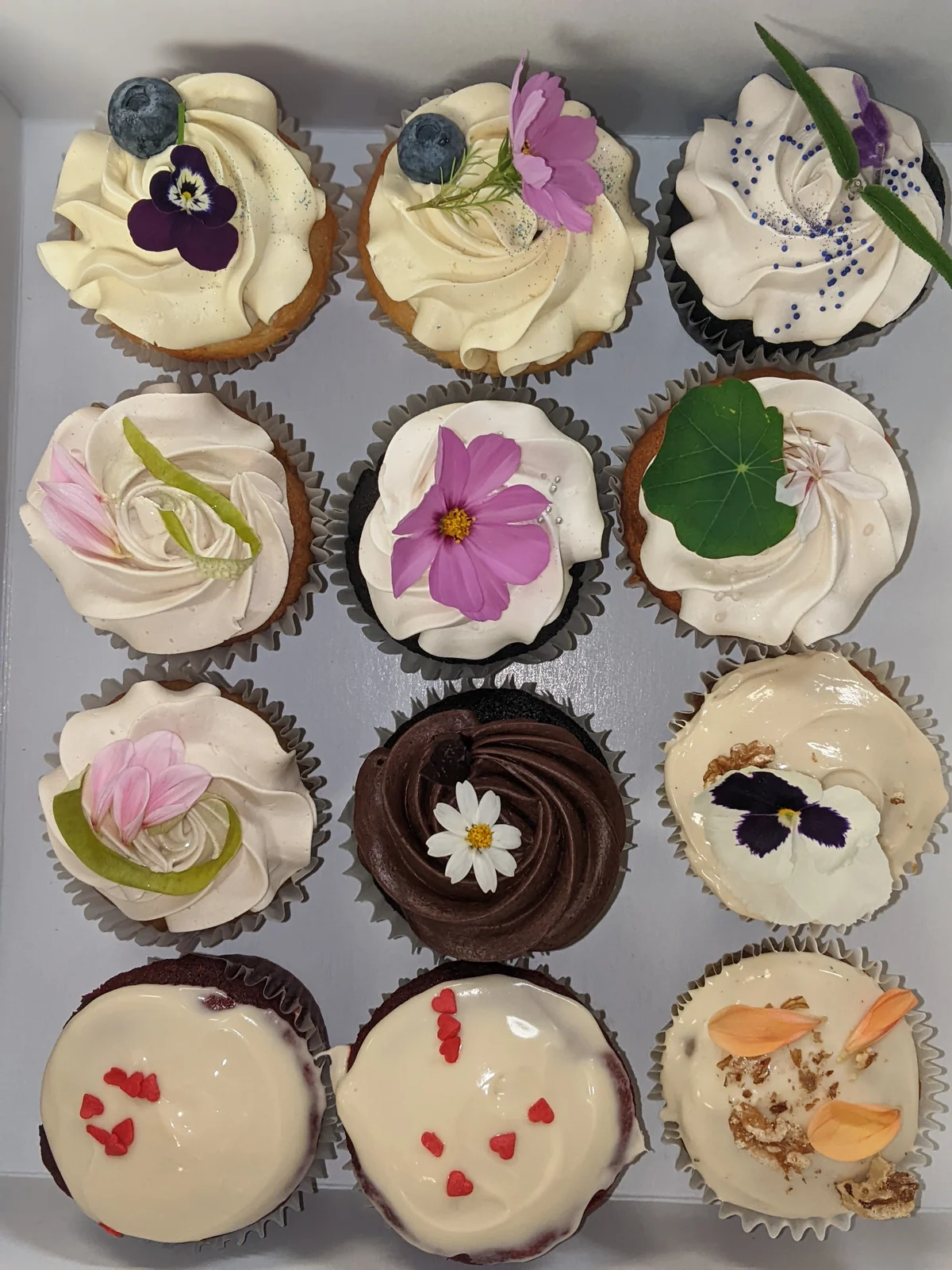 A dozen cupcakes in a box, each topped with a different edible flower