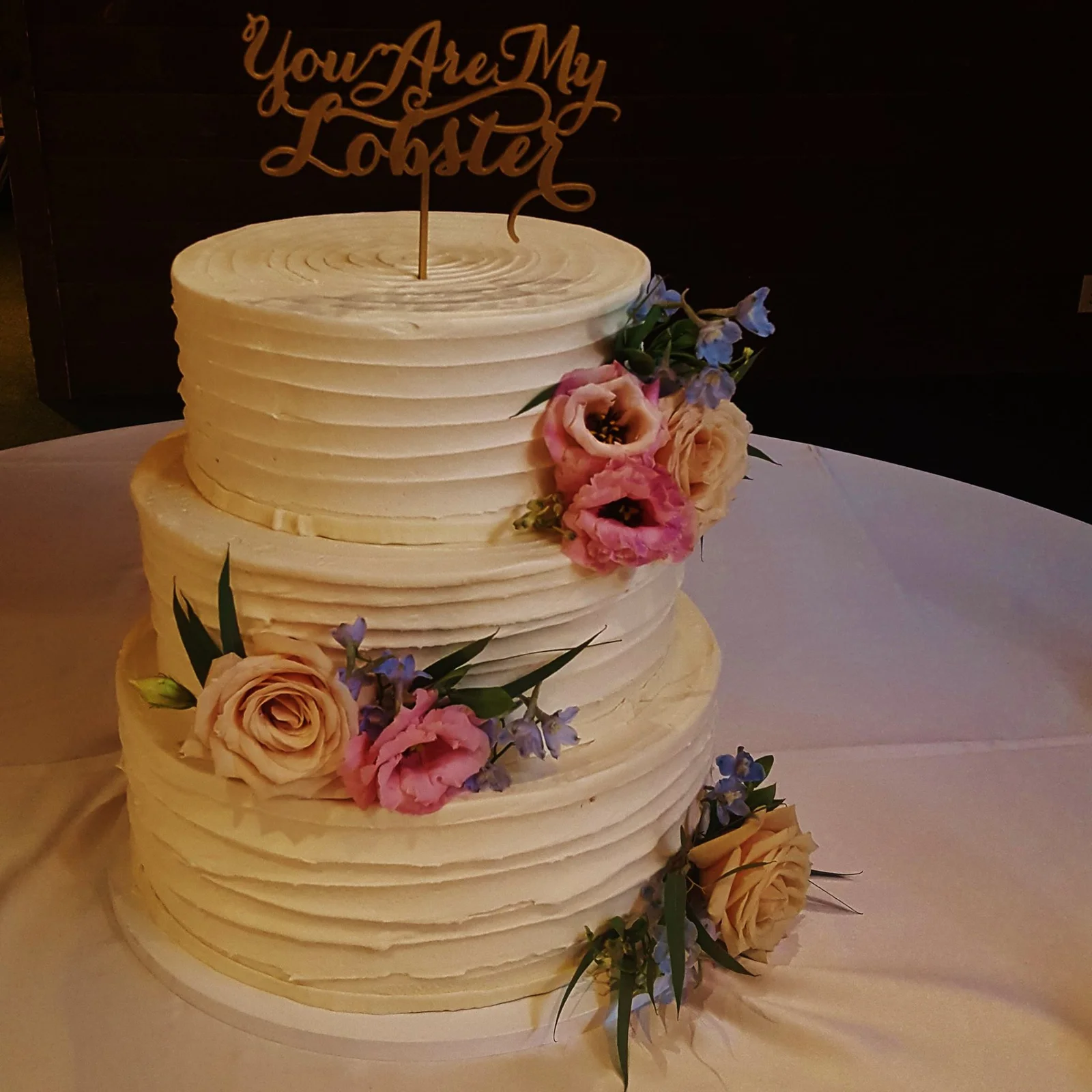 Three-tier white buttercream wedding cake with roses, lisianthus and delphinium on a reception table